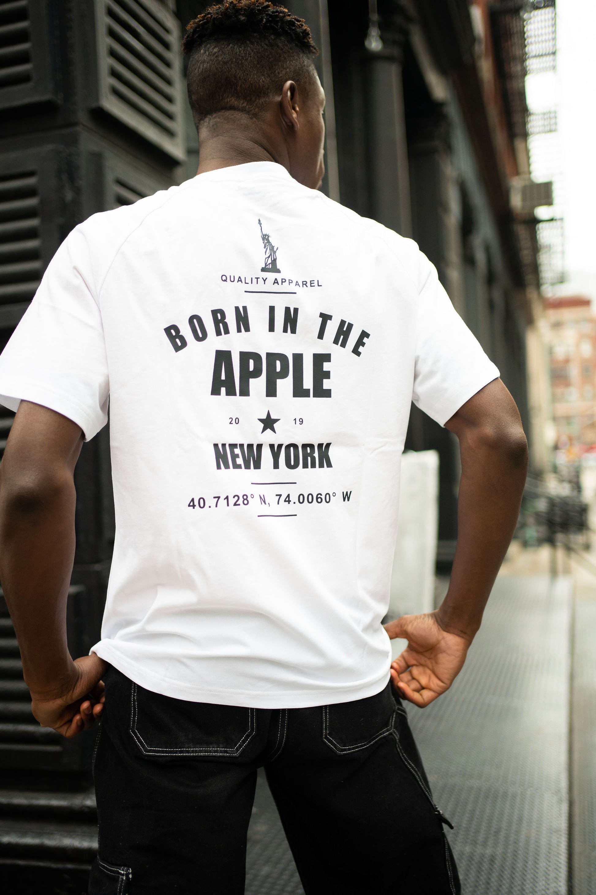 Oversized white t-shirt, heavyweight and 100% cotton. Black embroidered BITA Torch on left chest, grey jock tag with black born in the apple text on bottom left edge, and black screen printed Born In  The Apple text covers the back. Raglan sleeves. 